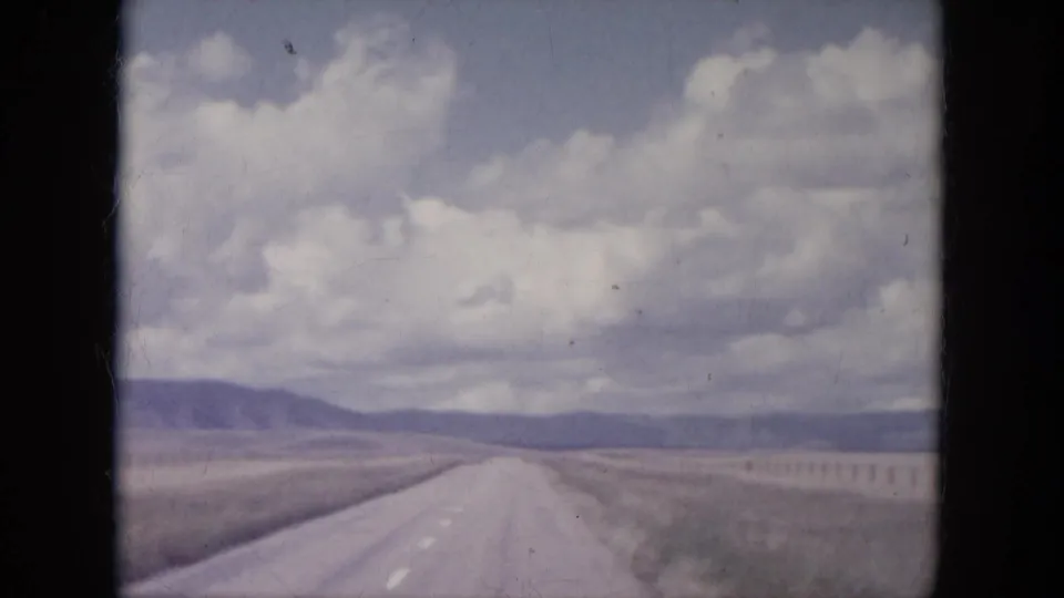 1966 South Dakota 8mm view of a straight road, fence line, open field, and distant hills under heavy clouds.