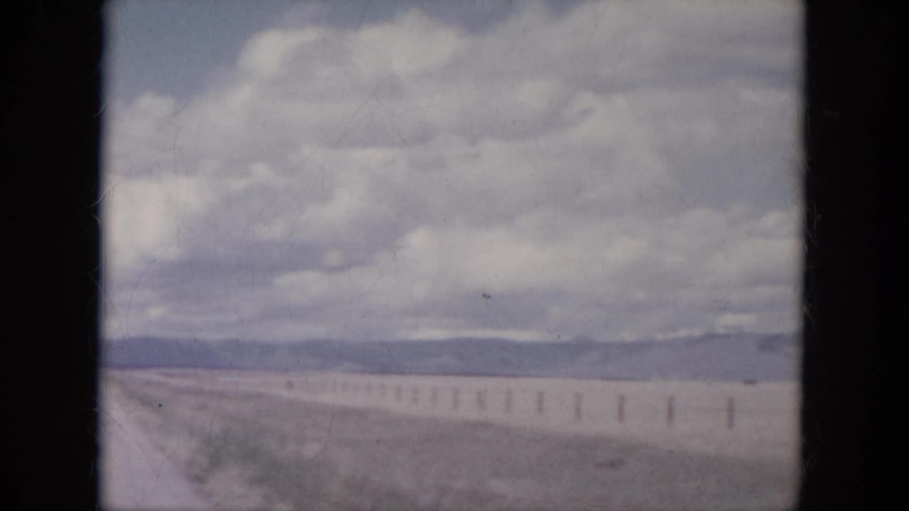 1966 South Dakota roadside view with fence posts, pale open land, distant low hills, and a clouded sky.