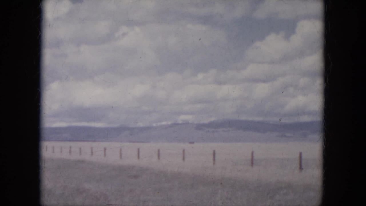 1966 South Dakota travel footage frame with fence posts crossing a broad plain below distant hills and cloud cover.