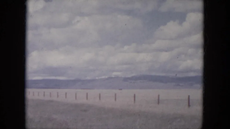 1966 South Dakota travel footage frame with fence posts crossing a broad plain below distant hills and cloud cover.