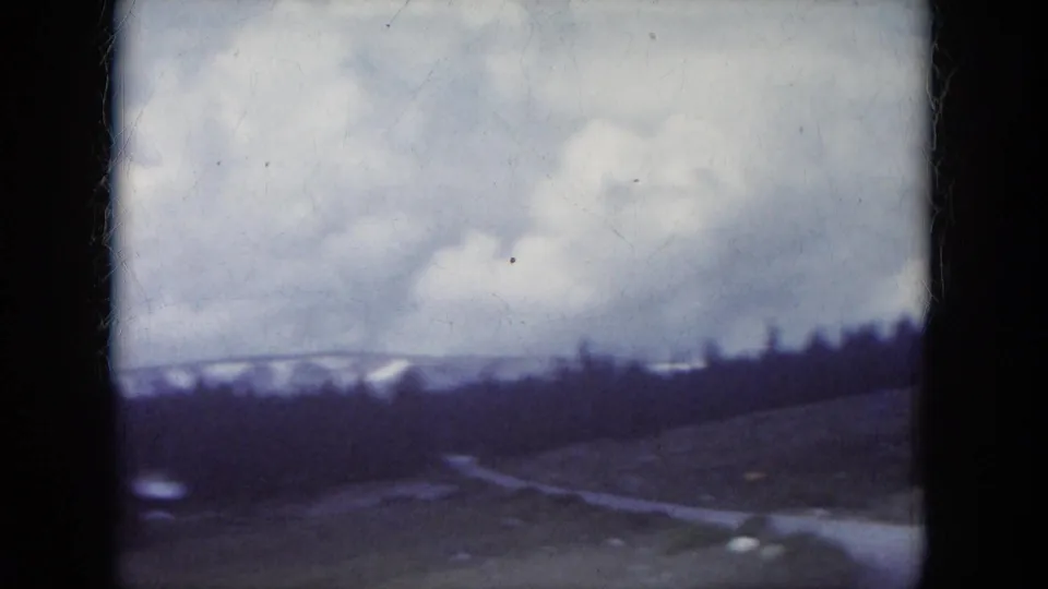 1966 South Dakota home-movie frame showing dark evergreen hills, a winding road, clouds, and distant snow patches.
