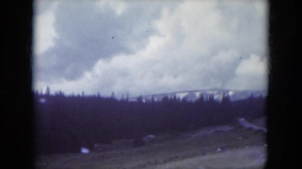 1966 South Dakota landscape frame showing forested hills, a pale road or clearing, distant snow, and large clouds.