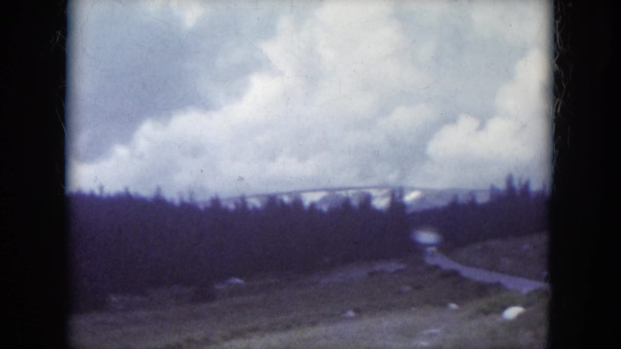 1966 South Dakota 8mm frame showing a winding road through dark pines, grassy roadside, rocks, clouds, and snow-dusted hills in the distance.