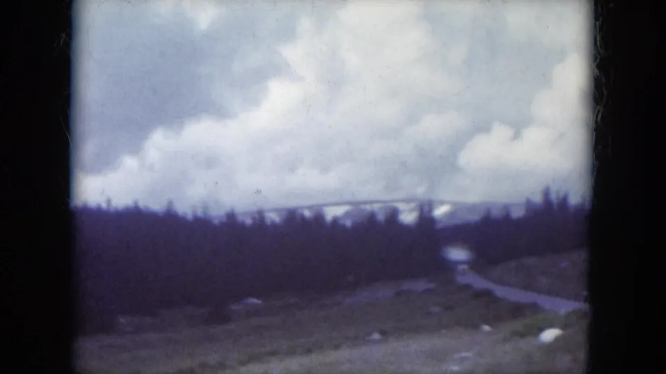 1966 South Dakota 8mm frame showing a winding road through dark pines, grassy roadside, rocks, clouds, and snow-dusted hills in the distance.