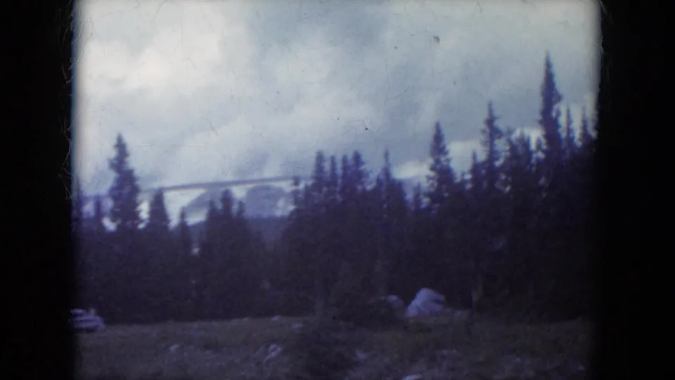 Soft 8mm frame of a South Dakota mountain roadside, with pine trees, a small rock outcrop, grass, cloudy sky, and snow on distant hills.
