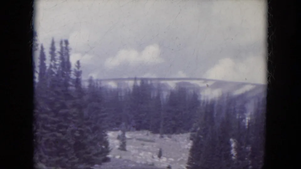 1966 South Dakota 8mm view across pine trees to an open mountain slope with scattered snow, pale clouds, and soft home-movie exposure.