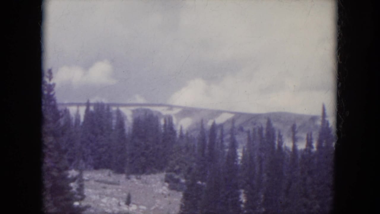 1966 South Dakota home-movie frame showing distant snowy mountain slopes and dense evergreens through soft 8mm color film.