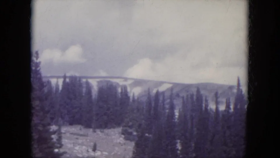 1966 South Dakota home-movie frame showing distant snowy mountain slopes and dense evergreens through soft 8mm color film.