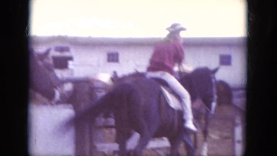 1966 South Dakota 8mm frame of a rider in a red shirt and cowboy hat on a dark horse near a simple ranch building.