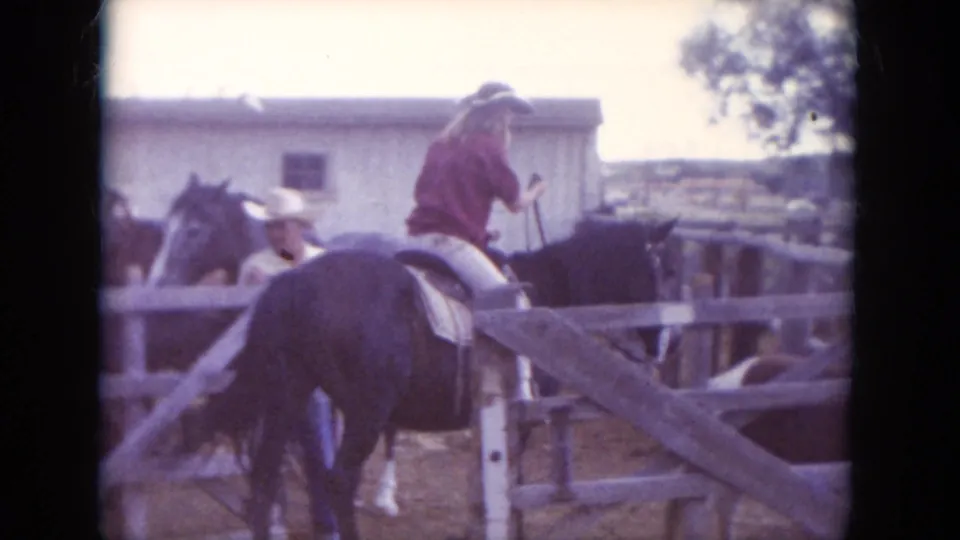1966 South Dakota home-movie frame showing a rider in a red shirt on a dark horse near wooden fencing and other horses.