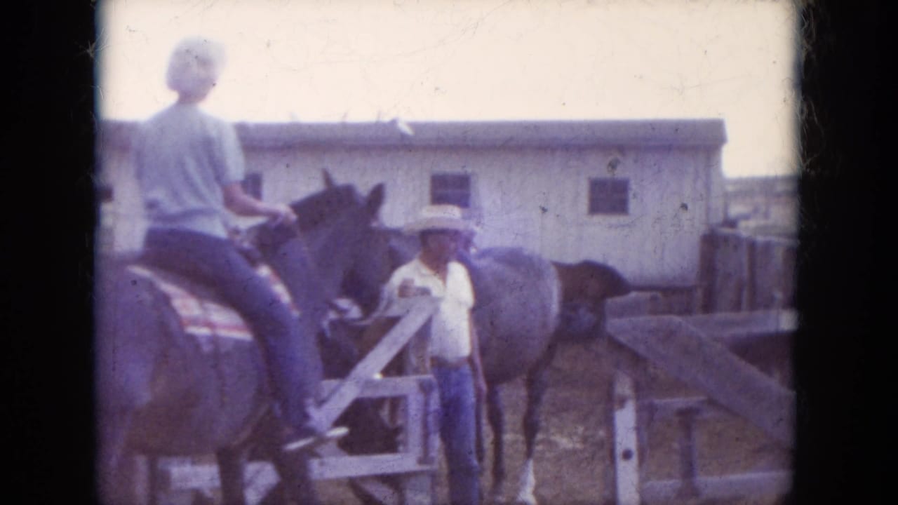 1966 South Dakota home-movie frame with a rider in a light shirt on horseback near a standing person, wooden corral fencing, and a pale barn.