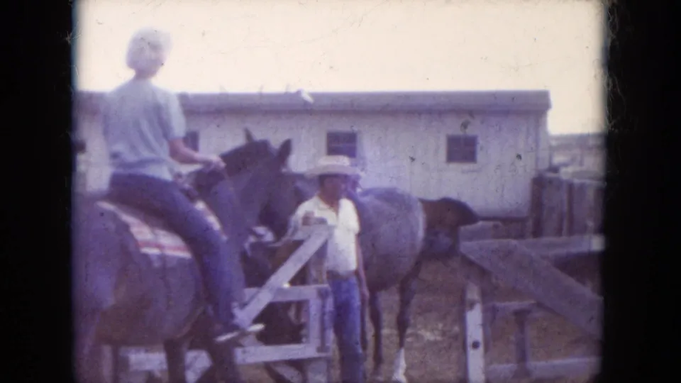 1966 South Dakota home-movie frame with a rider in a light shirt on horseback near a standing person, wooden corral fencing, and a pale barn.