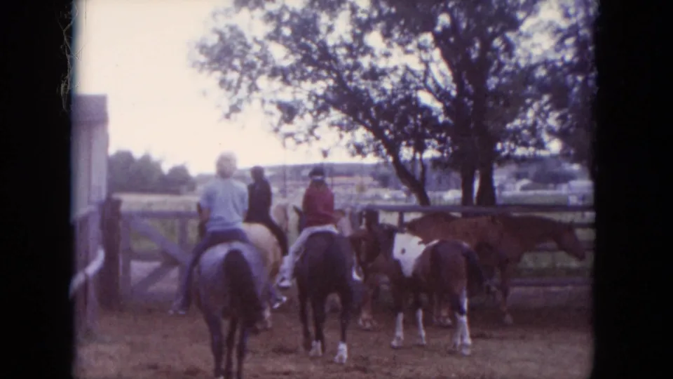1966 South Dakota home-movie frame showing multiple horseback riders moving through a fenced dirt arena beside a barn and large tree.