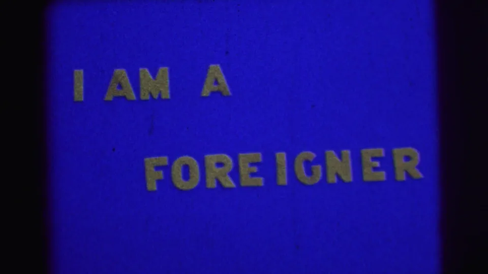 Static blue 8mm frame from 1967 Brazil with yellow block text reading I AM A FOREIGNER.