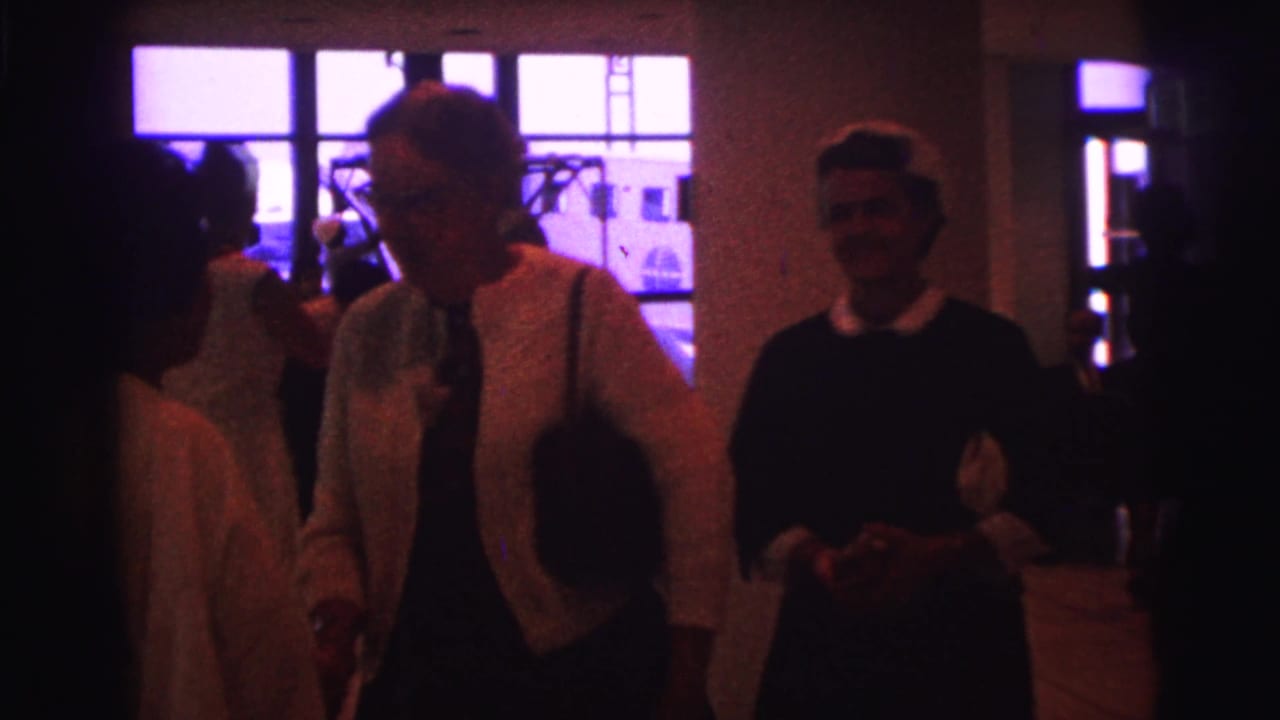 Adults in a dim 1967 Brazil interior gather near bright windows while women appear to exchange greetings.