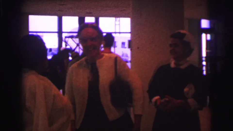 Women in period clothing stand in a softly lit Brazil interior, one smiling as a greeting continues.