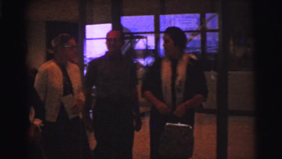 Three adults in a dim 1967 Brazil interior stand together near windows and a glass door after the greeting sequence.