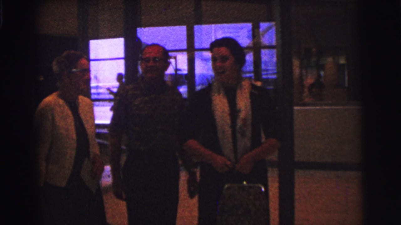 1967 Brazil home-movie frame showing three adults indoors, including a woman in a light cardigan, with blue reflections in large windows.