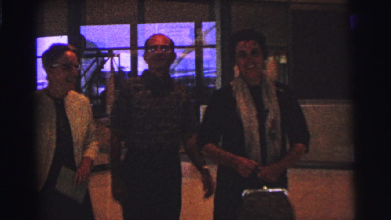 Three adults walk in front of large windows, with the central man facing the camera.