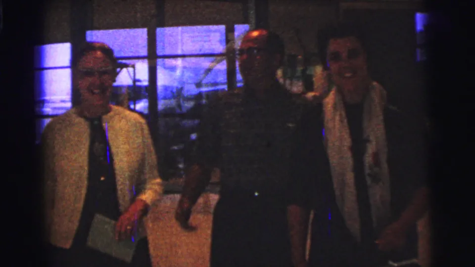 1967 Brazil home-movie frame showing a woman with glasses beside two men, moving through a dim interior with large windows.
