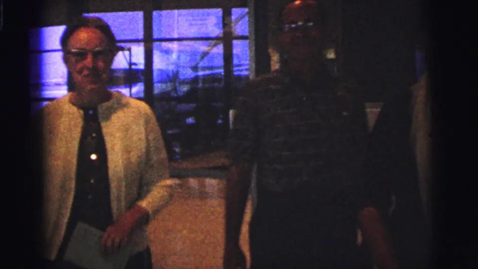 1967 Brazil frame showing a woman in a light cardigan and a man in a dark patterned shirt near large glass windows.
