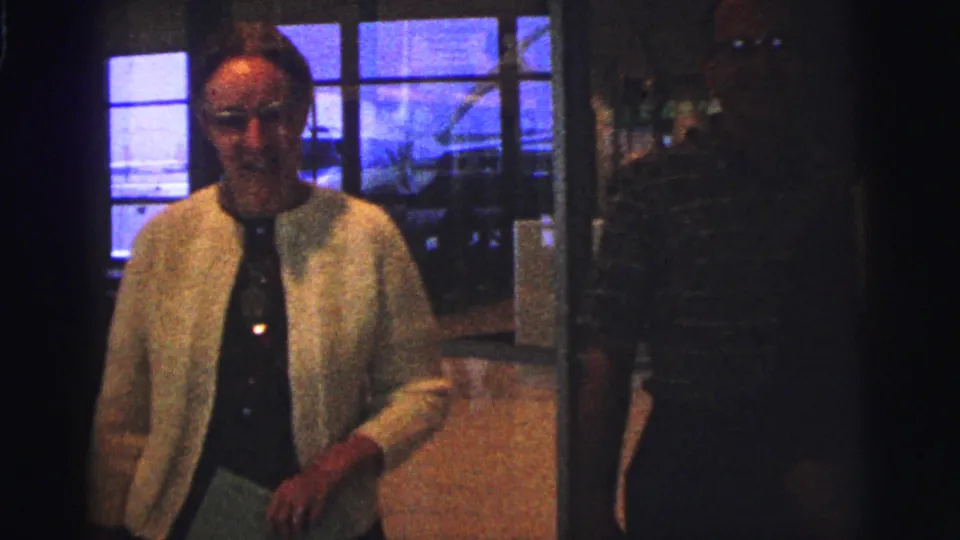 1967 Brazil home-movie frame showing a woman in a light cardigan moving through a dim airline-terminal interior.