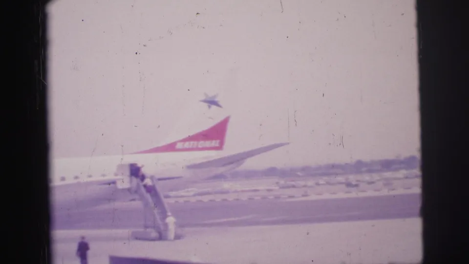1967 Brazil 8mm frame showing passengers boarding a National Airlines aircraft on mobile stairs under a hazy sky.