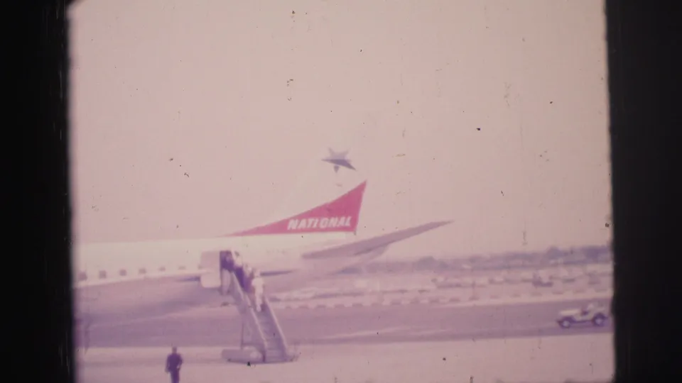 1967 Brazil airport frame with passengers on mobile stairs, a National Airlines tail, and a small vehicle on the apron.