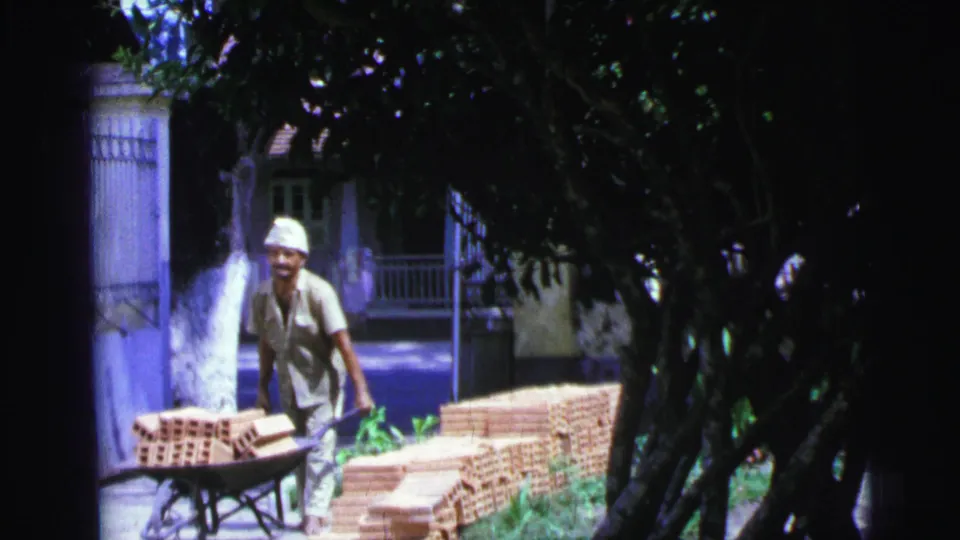 In 1967 Brazil home-movie footage, a worker in a white cap pushes red bricks in a wheelbarrow through a sunlit yard.