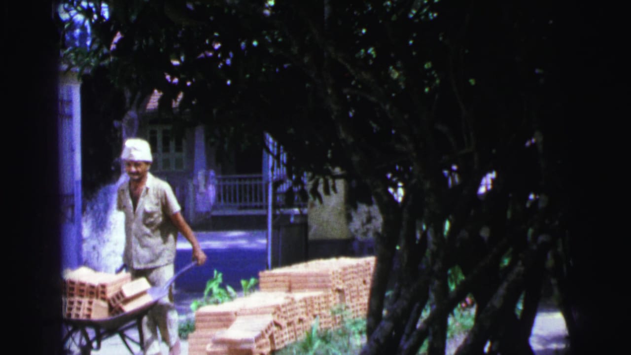 Worker continues pushing a brick-filled wheelbarrow near a gate and shaded trees.