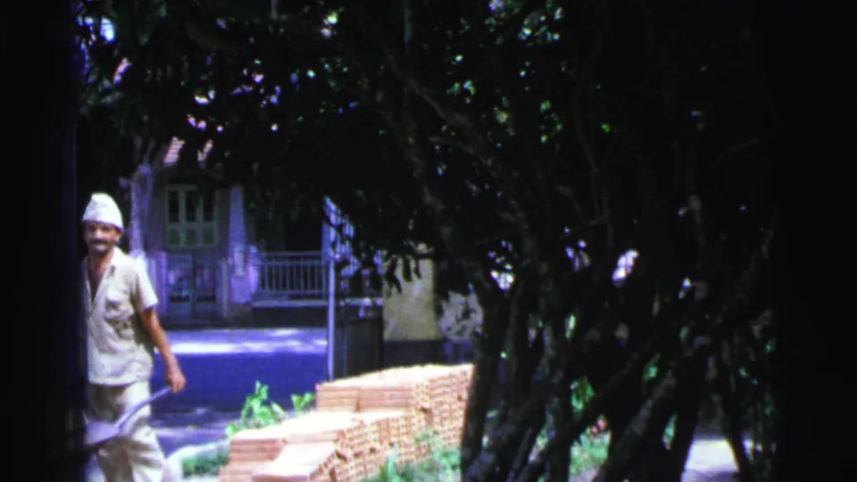 The worker is partly cut off at the left edge as he pushes the wheelbarrow past brick stacks in the shaded Brazil yard.