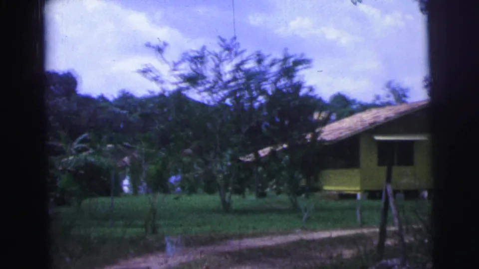 1967 Brazil home-movie frame showing a yellow rural house with a tiled roof set behind grass, trees, fence posts, and a dirt track.
