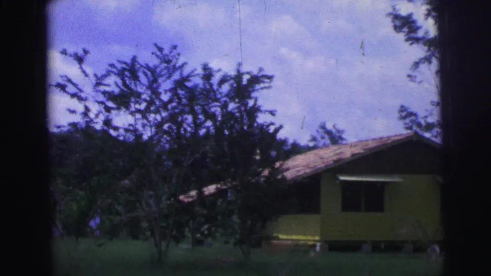 1967 Brazil frame with a closer view of the yellow house, tiled roof, dark window, trees, grass, and blue-shifted cloudy sky.