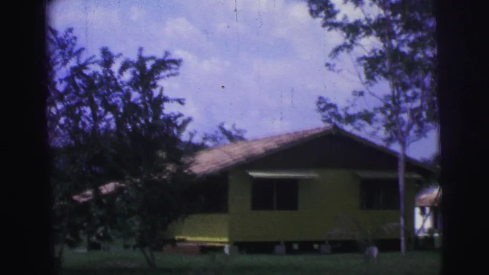 1967 Brazil 8mm frame showing a yellow rural house behind trees, with grass and a cloudy sky.
