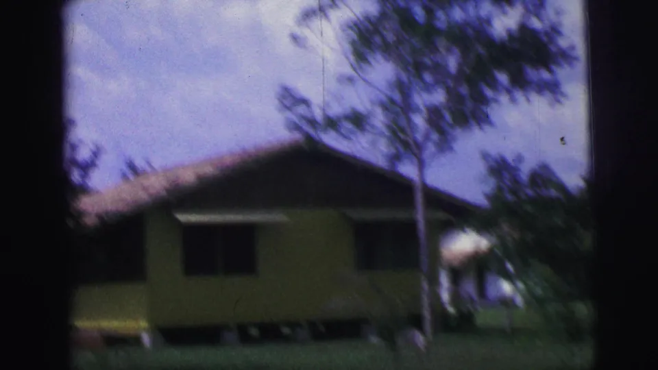 Soft 8mm view of a yellow house in rural Brazil, partly obscured by foreground trees and motion blur.