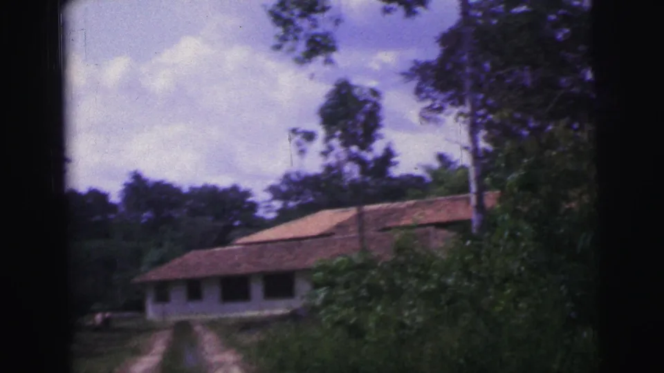 1967 Brazil 8mm frame approaching a long single-story house with a red tiled roof, shaded windows, and surrounding trees.