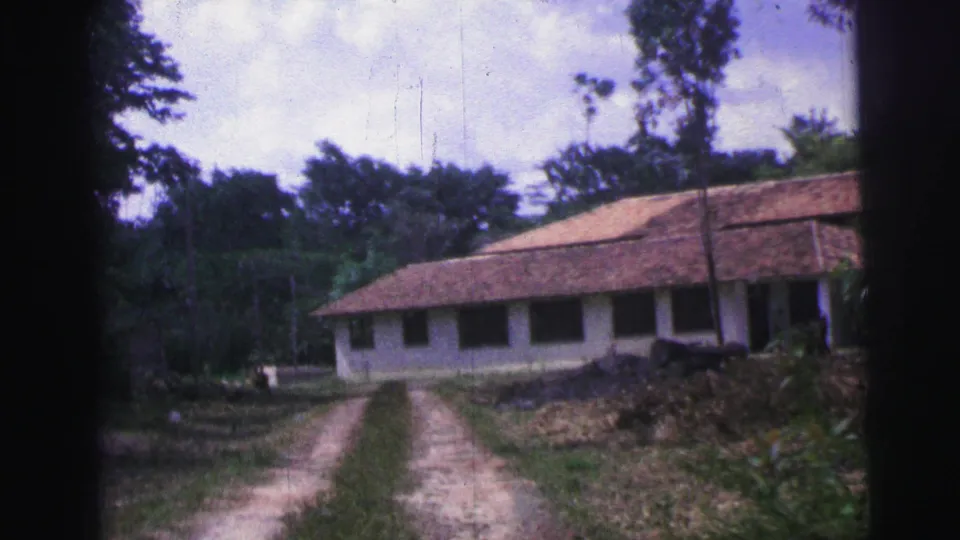 1967 Brazil 8mm frame showing a closer exterior view of a long house with shaded windows, tiled roof, dirt track, and trees.