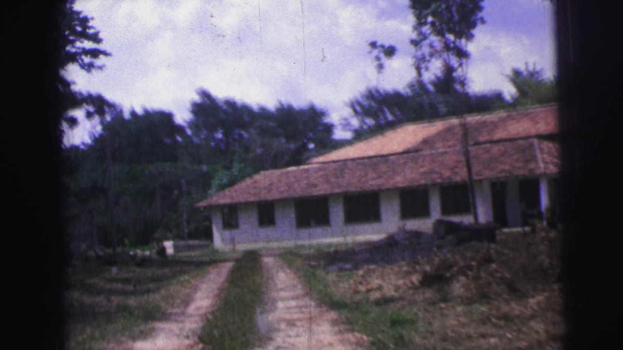 Dirt track leading toward a long white building with a tiled roof among trees.