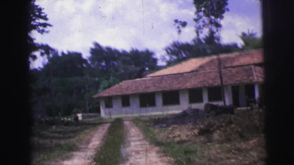 1967 Brazil 8mm frame showing a dirt drive approaching a long white building with dark windows, terracotta roof tiles, and trees around it.