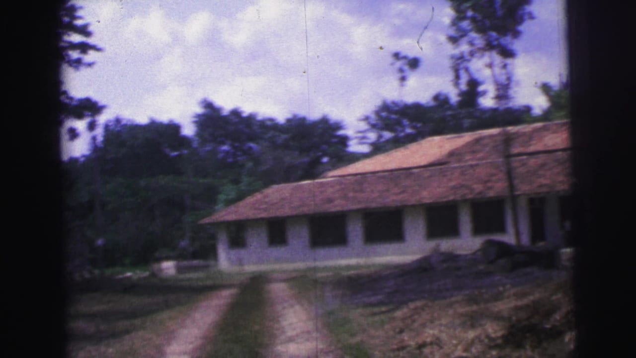 1967 Brazil 8mm frame from a moving approach toward a long white building with a terracotta roof, dark windows, and wooded surroundings.