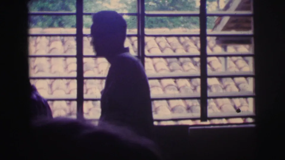 Interior 1967 Brazil 8mm frame with a silhouetted person beside a barred window, roof tiles visible beyond the bright opening.