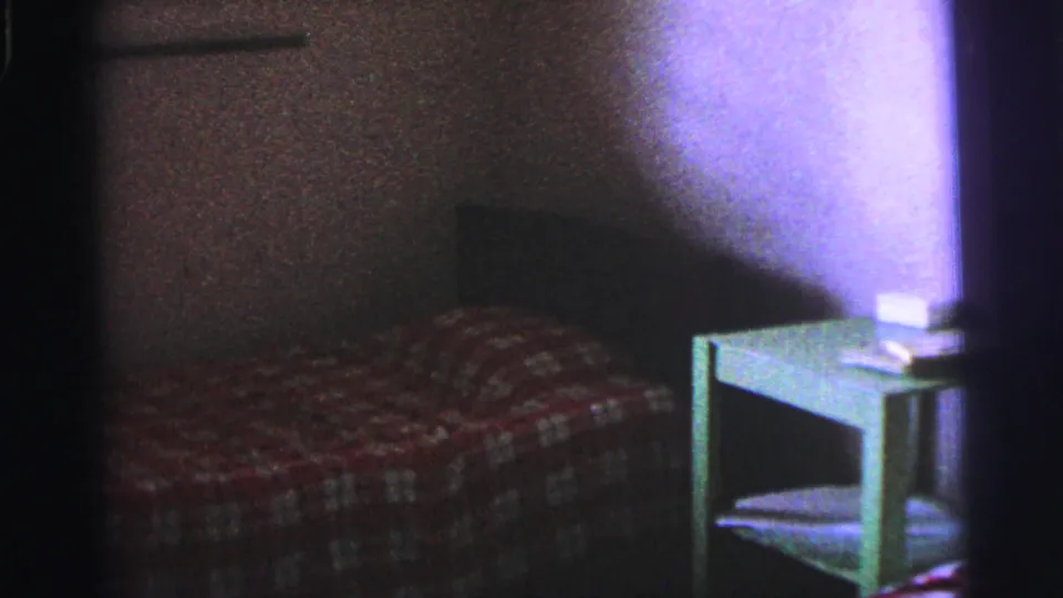 1967 Brazil 8mm frame of a sparse room with a checked bedcover, wall shadow, and small green bedside table.