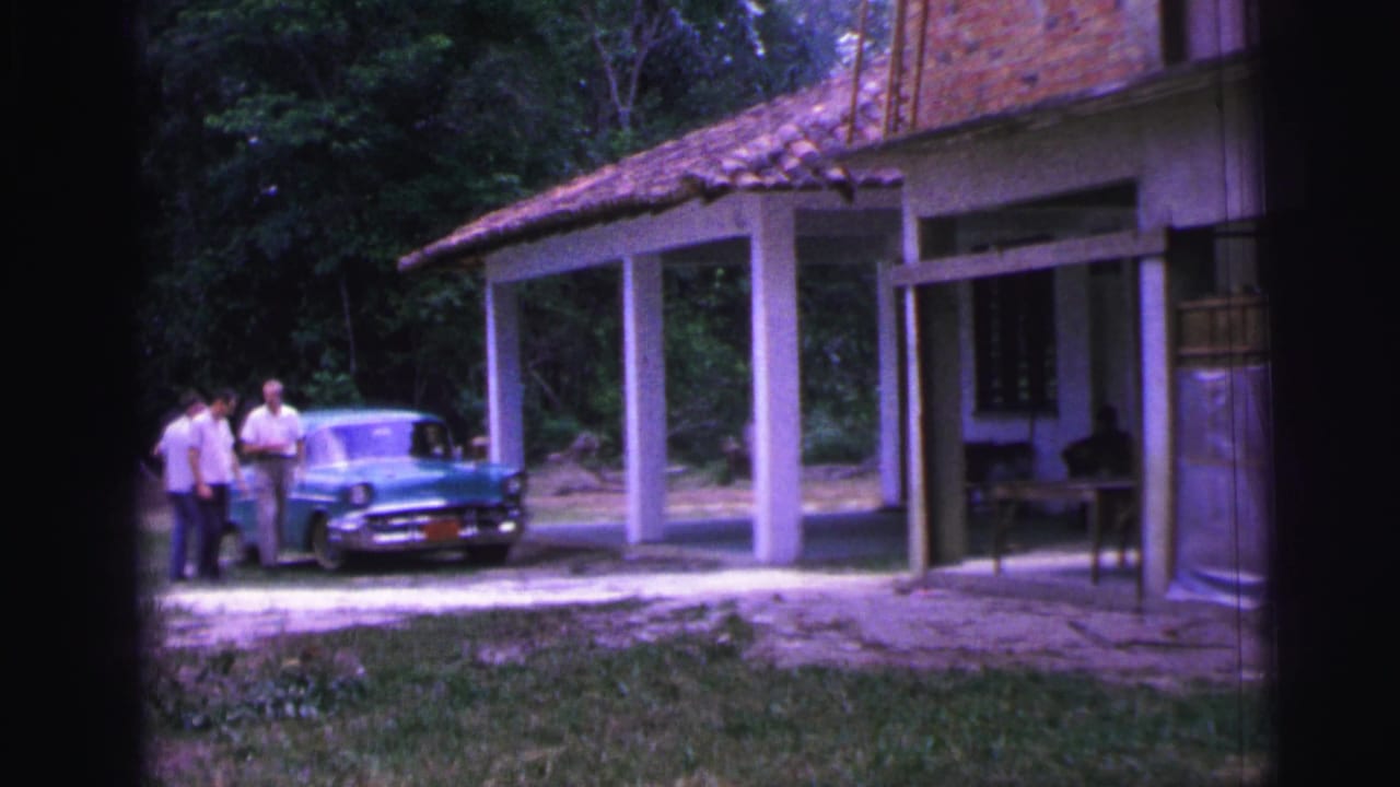 1967 Brazil home-movie frame showing two men near a turquoise sedan in front of a brick villa with columns and trees.