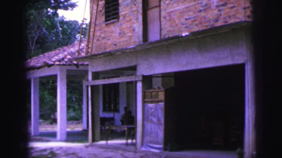 1967 Brazil home-movie view of a brick villa exterior with tiled porch roof, white columns, and a person seated in shade.