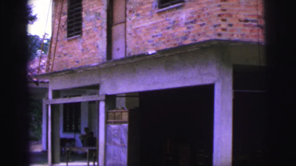 1967 Brazil frame showing the villa facade closer, with brick walls, porch columns, an open dark doorway, and shaded seating.