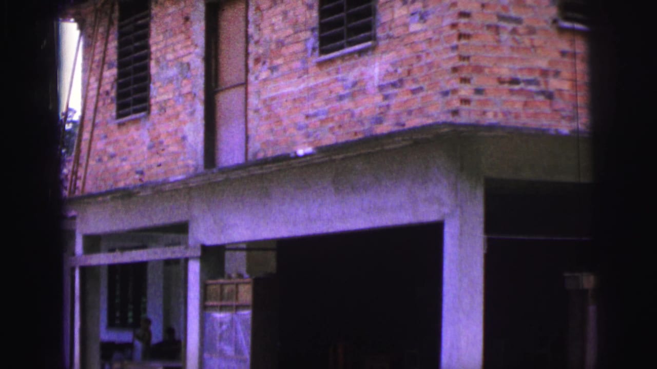 1967 Brazil 8mm frame of a weathered brick house with shuttered windows, concrete supports, and a shadowed open lower level.