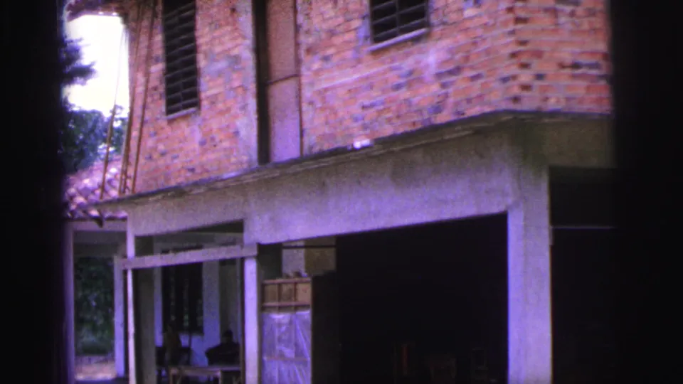 The camera holds on the brick house exterior in Brazil, with concrete supports, upper shutters, and a dark open bay below.