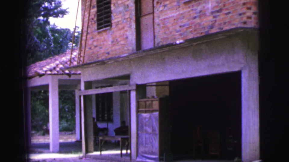 1967 Brazil home-movie frame showing a brick house, open lower level, concrete posts, roof tiles, and indistinct items in the shaded interior.
