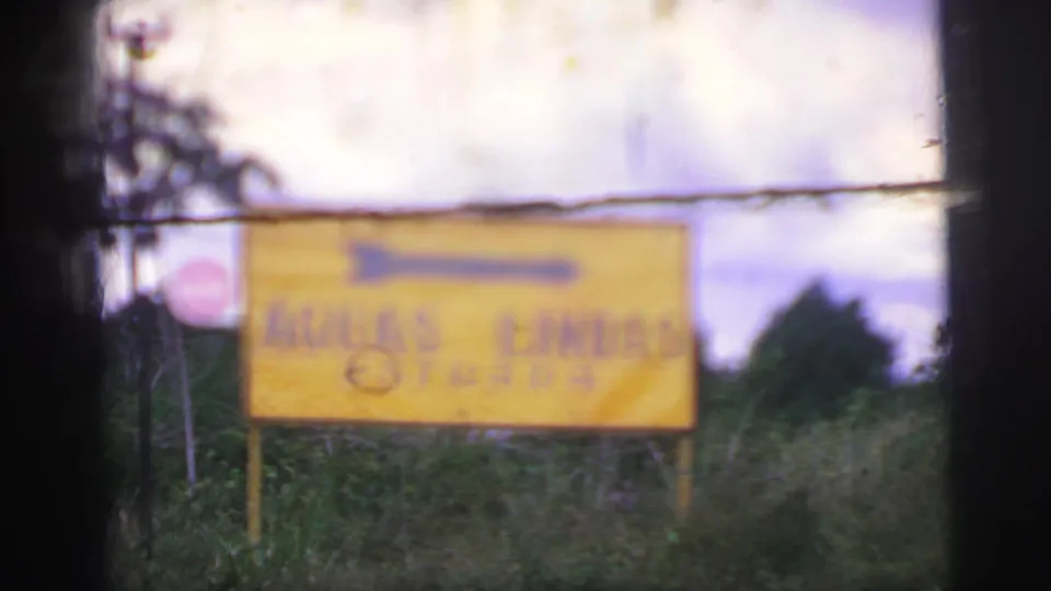1967 Brazil 8mm frame of a yellow Águas Lindas Entrada road sign with a directional arrow against roadside greenery and cloudy sky.
