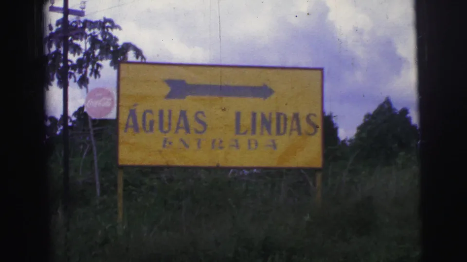 1967 Brazilian home-movie frame with an Águas Lindas entrance sign and right arrow set against grass, trees, and clouds.